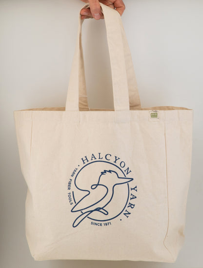 The Halcyon Yarn Recycled Cotton Tote Bag w/Pocket, a beige tote crafted from recycled cotton canvas, proudly displays a simplistic blue bird logo along with the text "Halcyon Yarn" in a circular design that reads "Since 1971" and "Fine Fiber Tools." Hanging gracefully from one's hand, an "ECO" tag is visible on the side, making this oversized canvas tote both stylish and sustainable.