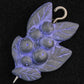 The Woodland Shawl Pin by Bonnie Bishoff Designs, crafted by Bonnie Bishoff, features a decorative design resembling a cluster of berries and leaves. Made from polymer clay with a predominantly purple metallic sheen, it is lightweight and equipped with a hook and pointed end for securing to shawl closures. The solid black background accentuates the intricate details of this beautifully designed pin.