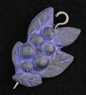 The Woodland Shawl Pin by Bonnie Bishoff Designs, crafted by Bonnie Bishoff, features a decorative design resembling a cluster of berries and leaves. Made from polymer clay with a predominantly purple metallic sheen, it is lightweight and equipped with a hook and pointed end for securing to shawl closures. The solid black background accentuates the intricate details of this beautifully designed pin.