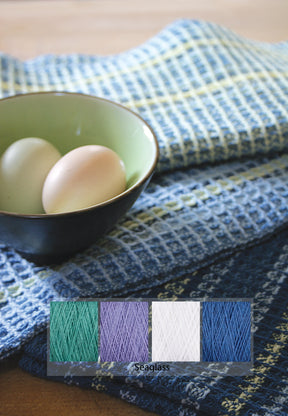 A bowl with two eggs sits on a table covered with the Waffle Weave Dish Towel Kit from Halcyon Yarn. Below the bowl rests a color palette featuring four yarn colors labeled "Seaglass" in green, lavender, white, and blue shades—perfect for your next weaving kit project.