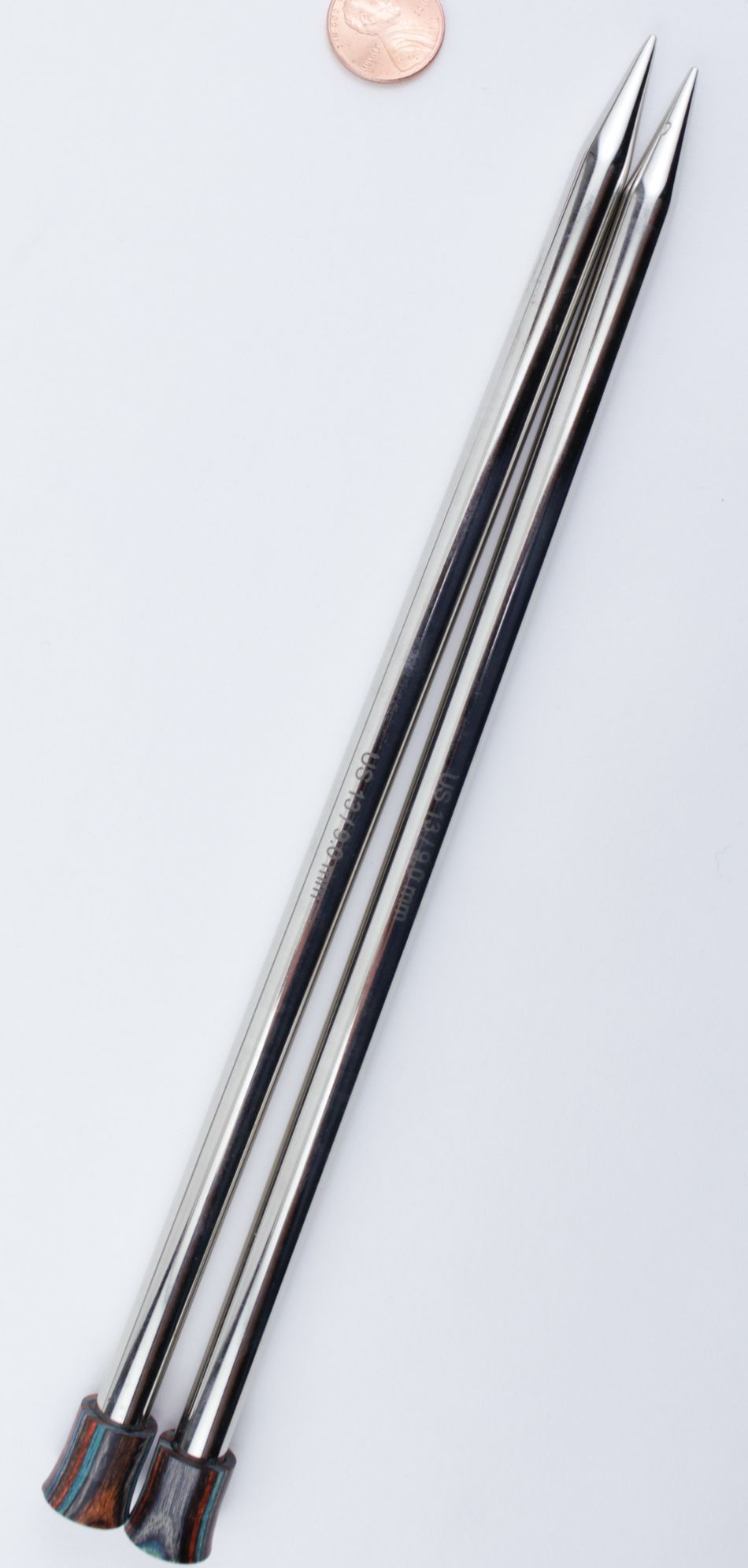 Two metal knitting needles from the Accessories Unlimited Nova Platina Single Point Knitting Needles collection lie parallel to each other on a white surface. The straight needles, with sharp pointed ends and dark-colored caps, have a penny placed near the tips for scale.