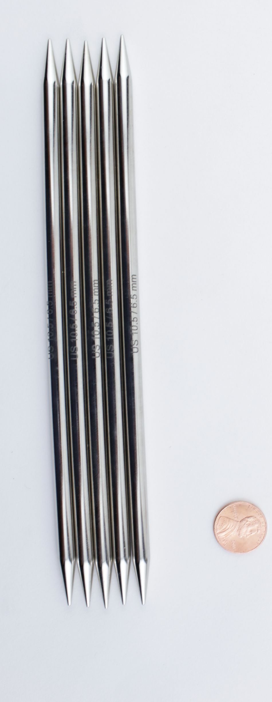 Six Accessories Unlimited Nova Platina Double-Point Knitting Needles, crafted from high-quality chrome, are arranged parallel to each other with their tips aligned at both ends. They are placed next to a single penny for size reference on a white background.