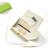 An open green case from the Skacel Addi-Click Interchangeable Needle Set reveals a set of neatly arranged knitting needles with bamboo tips. Around the case lies an Addi cord with wooden tips. In the top-left corner, there's an inset showing the closed case. The interior of the case features a patterned lining.