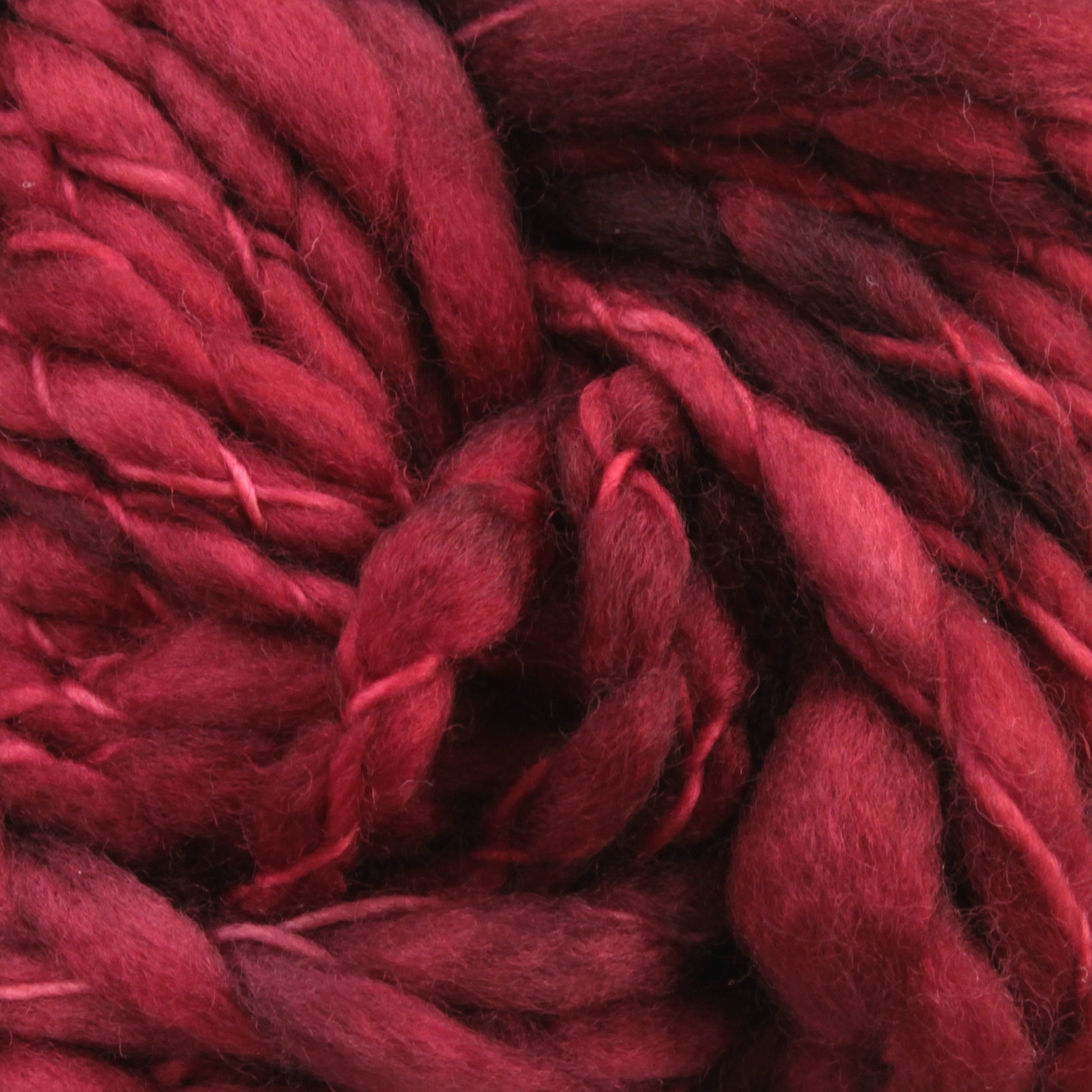 Close-up of Malabrigo Caracol by Malabrigo Yarn, showcasing red-hued roving wool fibers with a lush, textured pattern. The strands are variably thick and twisted together, resembling super bulky yarn with a mix of deep crimson and lighter red tones. The fibers appear soft and fluffy, ideal for knitting or other fiber crafts.