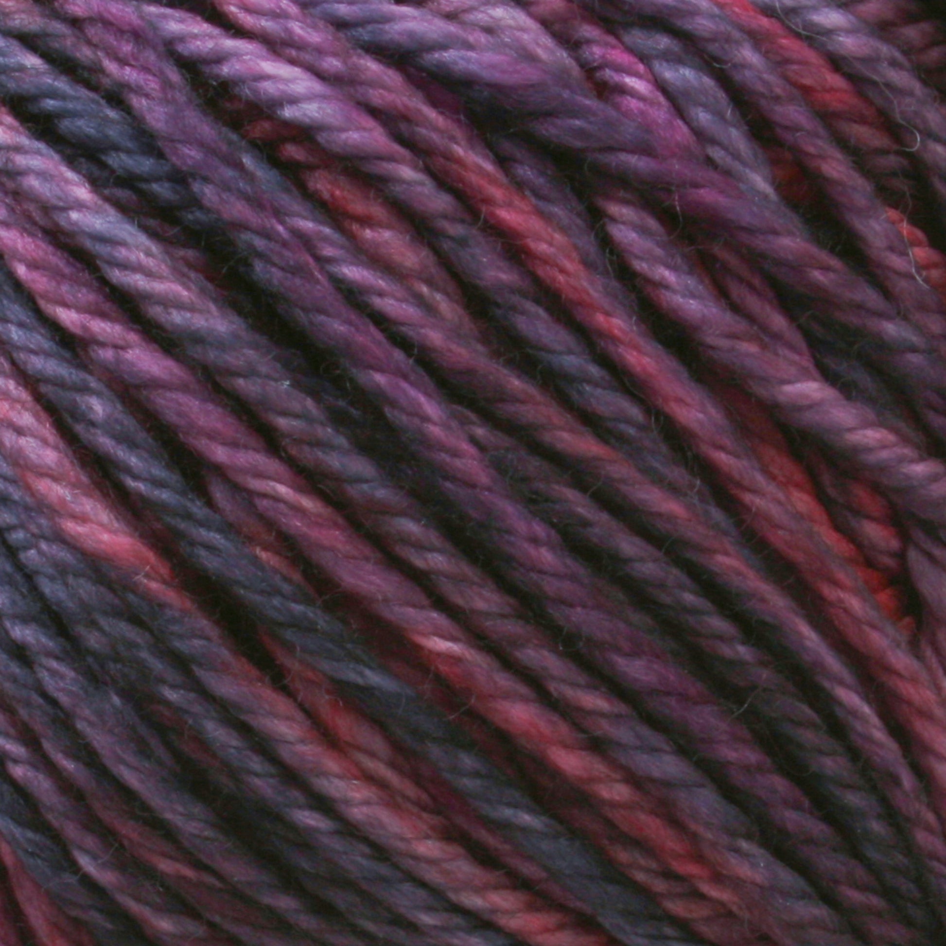 Close-up image of multiple strands of yarn intertwined, showcasing a blend of deep purple, magenta, and hints of blue hues. The texture appears soft and slightly shiny, indicating its suitability for knitting or crocheting. The kettle-dyed colors accentuate the luxurious feel of Malabrigo Yarn's Rios yarn.