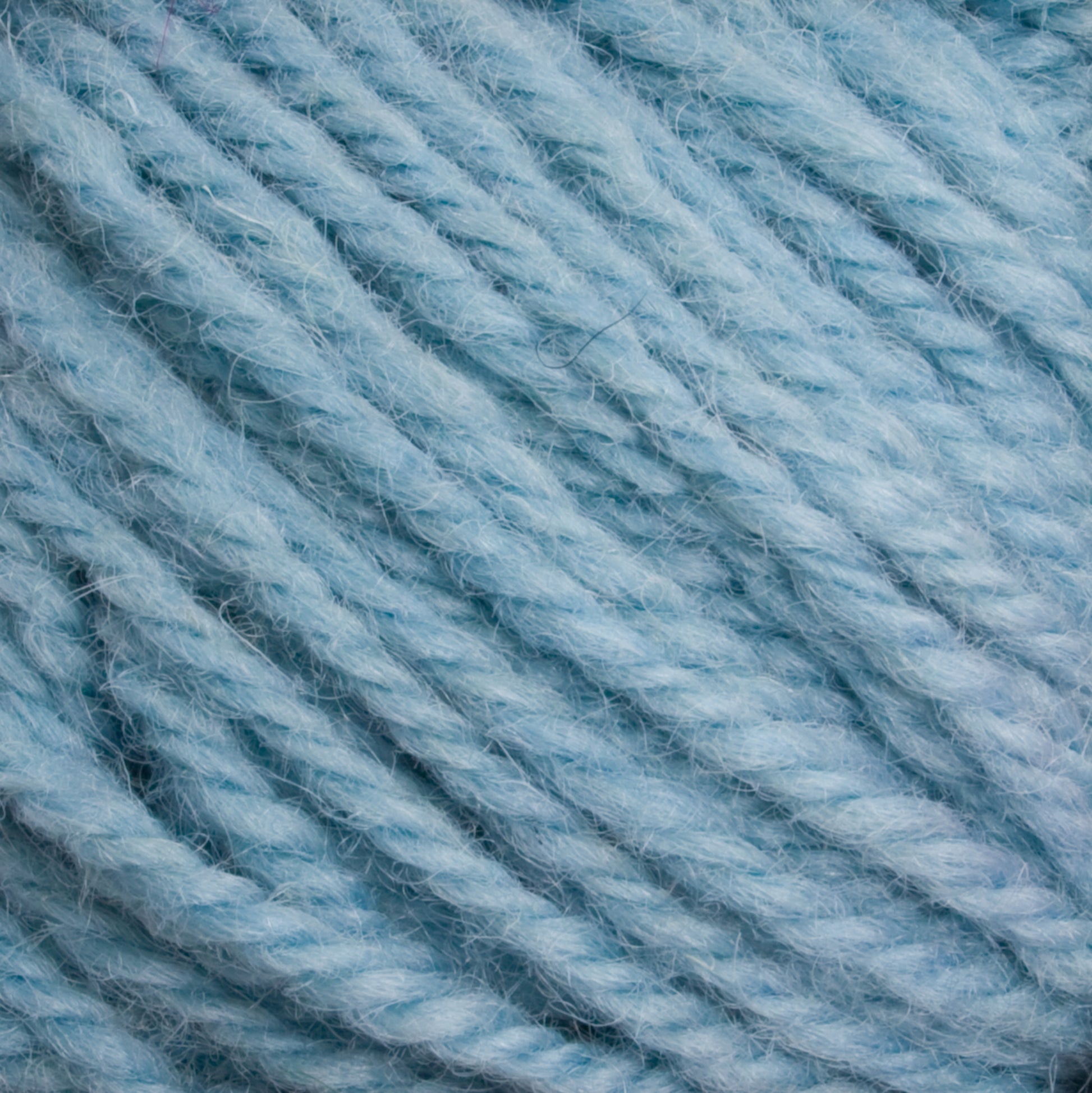 Close-up of Halcyon Deco Rug Wool in light blue. The thick, slightly twisted strands create a textured surface. Made by Caledonian Dye Works from 100% wool, the material appears soft and fuzzy, making it ideal for knitting or crocheting cozy garments and accessories.