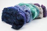 A close-up image showcasing a vibrant arrangement of Harrisville Fiber Batts Collection by Halcyon Yarn, featuring various colors including dark blue, purple, green, and light gray. The textured wool fibers are beautifully displayed on a plain white background, making them an excellent choice for felting projects or a colorful addition to your multi-color bags collection.