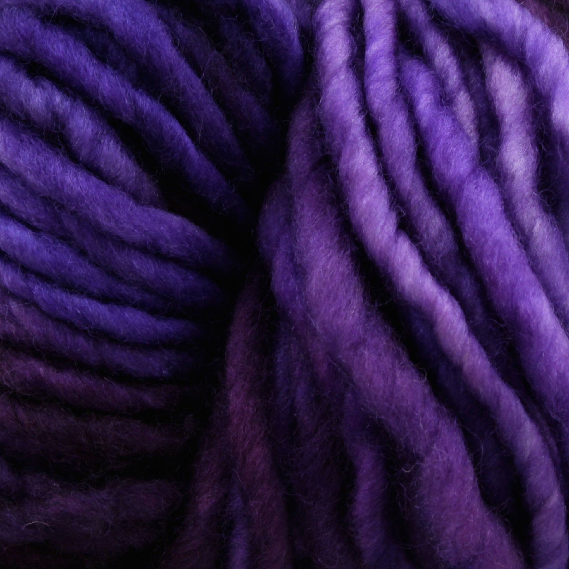 Close-up of fluffy, thick purple yarn with varying shades of purple intertwined. The texture is soft and woolly, showcasing detailed twists and loops in the strands. This Malabrigo Mecha superwash bulky yarn by Malabrigo Yarn is kettle dyed to perfection, adding depth and richness to every fiber.