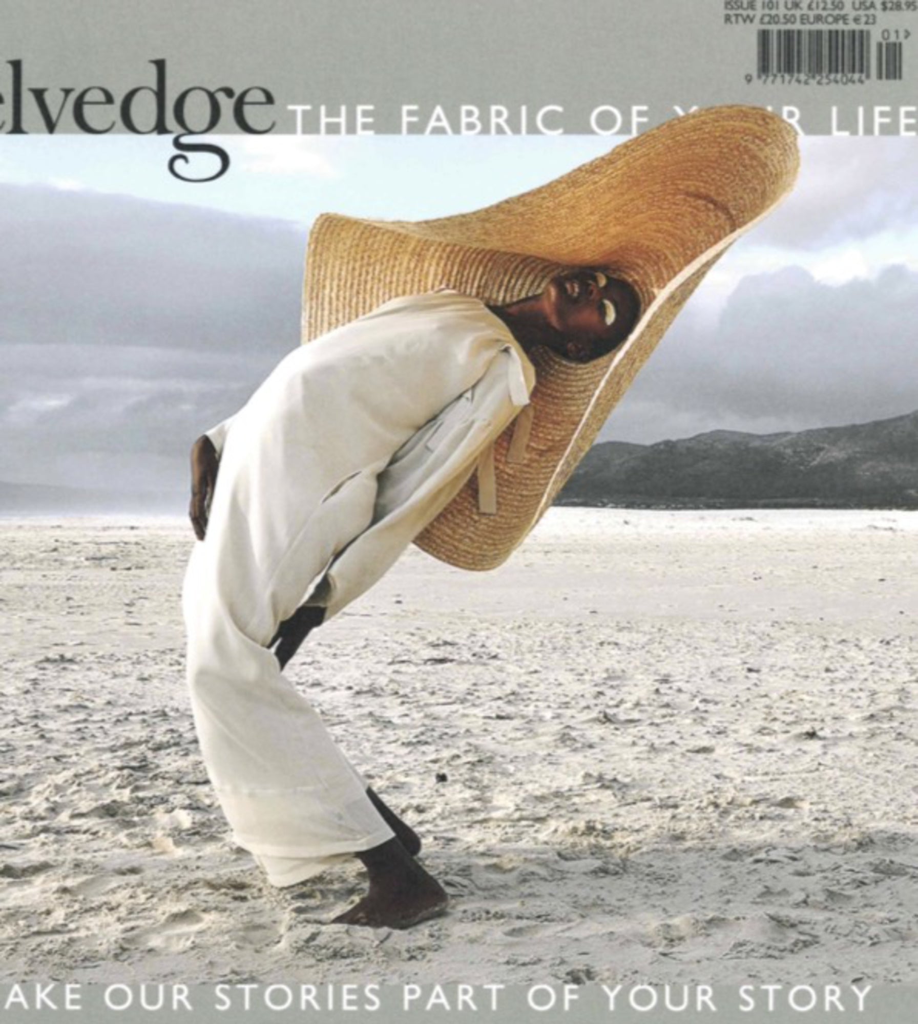 A person standing on a sandy beach wears a large, exaggerated straw hat and a loose, flowing white garment. They lean dramatically to one side, framed by a serene ocean and distant mountains in the background. The text "Selvedge - Issue 101: Grow" appears above.