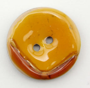 The Coco Boomerang 1 1/8" Button by Buttons Etc. features a glossy, round surface in shades of yellow and brown. It has two central sewing holes, with the top half predominantly yellow and the bottom displaying a brownish tint, creating an uneven color pattern reminiscent of shellac-coated buttons.