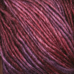 Close-up of Malabrigo Silky Merino yarn strands in mauve and burgundy intertwined, showcasing the texture and slight sheen of the fibers. The colors blend subtly from purple to red, highlighting the softness and complexity of this luxury yarn's hue and structure by Malabrigo Yarn.