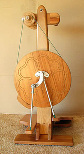 The Majacraft Suzie Spinning Wheel by Majacraft Co. is a vertically-mounted wooden spinning wheel with a modern design, featuring a circular, green lacquered wheel adorned with decorative engravings. It is mounted on a sturdy base with pedals, and includes a large wooden arm that extends upward to a small wheel at the top, ensuring smoother spinning action.