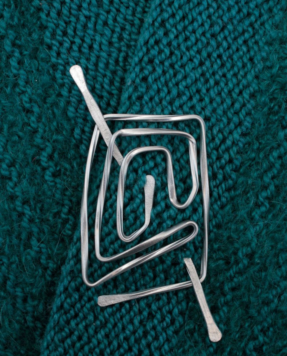 A tangled aluminum shawl pin from Carolyn Jackson, featuring an unusual and abstract shape, rests on a textured dark teal fabric background. The detailed weaving of the fabric is clearly visible, enhancing the contrast between the smooth metal and the soft textile.