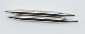 Two double-pointed knitting needles from the Accessories Unlimited Nova Platina Interchangeable Knitting Needles collection, sized "US 13 / 9.0 mm" with 4.5" tips, lie parallel on a plain white background like hollow brass pipes. One needle is positioned above the other, with the tips pointing in opposite directions.