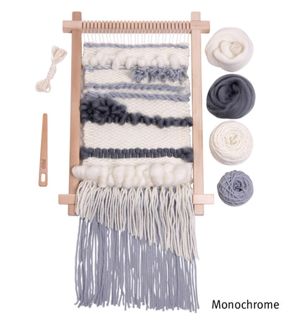 The Ashford Weaving Starter Kit from Ashford Handicrafts Limited features a wooden weaving loom working on a monochrome project in shades of white, gray, and black. Surrounding the loom are various coordinating balls of yarn, a wooden needle, and a bundle of wool. The word "Monochrome" is prominently displayed on this beautiful handwoven wall hanging.