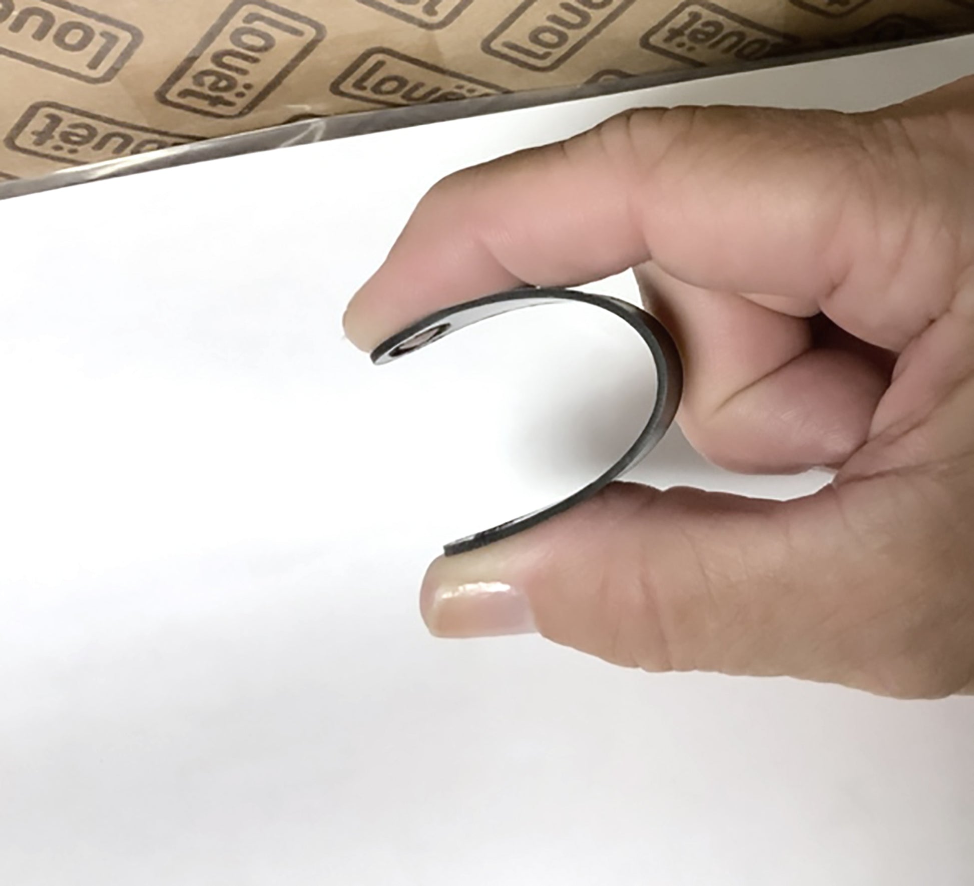 A person is holding a Louët Triangle Main Axel, a black, C-shaped object, between their thumb and index finger. The background shows a partially visible cardboard box with "loupe1" printed repeatedly on it. The image appears to focus on the object being held, possibly essential for repairing or maintaining Louët wheels.