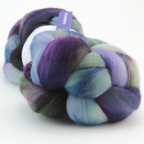 A close-up image of a skein of Nube by Malabrigo. The roving wool is thick and braided, showcasing a blend of colors including purple, teal, green, and white. A small, partially visible label reads Malabrigo Yarn attached to the skein. The background is plain and white.