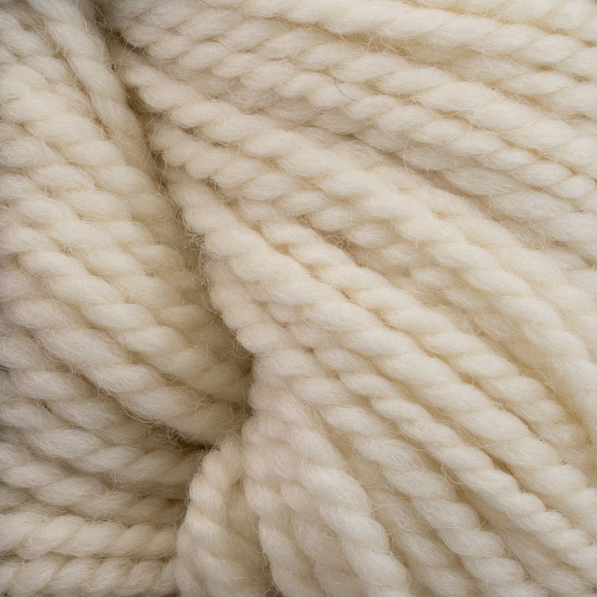 Close-up of thick, cream-colored Norumbega yarn by Caledonian Dye Works, showcasing the textured, twisted strands woven together. Made from 100% U.S. wool, the material appears soft and fluffy with a subtle sheen, creating a cozy and warm impression.