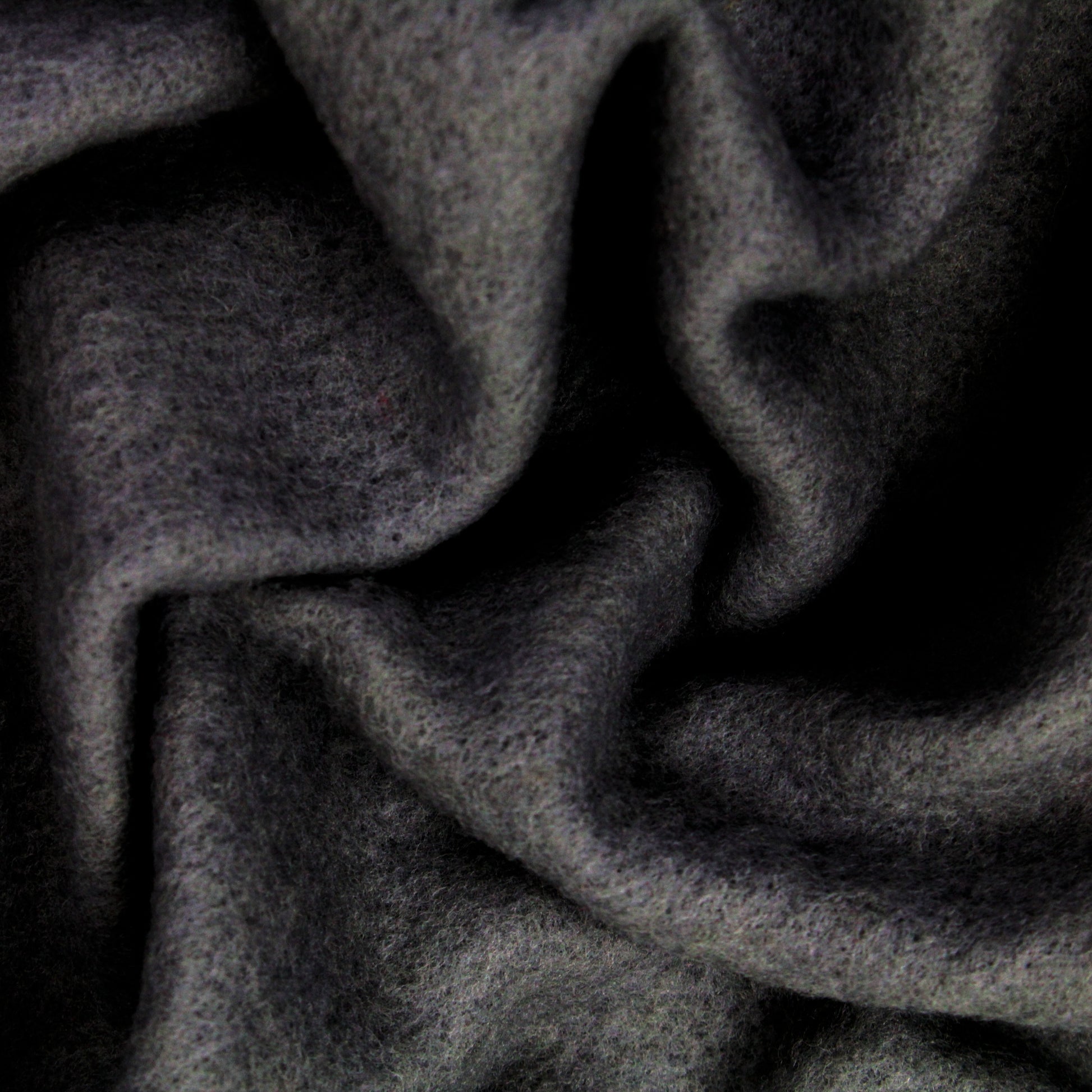 Close-up of Europa Wools Ltd's Merino Prefelt in dark grey, showcasing a soft, fuzzy texture that is ideal for felting projects or nuno felting. The material is folded and draped, forming gentle, undulating curves and shadows. Its surface appears smooth and plush to the touch.