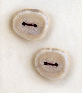 Two handcrafted Buttons - Antler from Favour Valley Woodworking lie on a beige background. The buttons are irregularly round and crafted from antler, with a smooth yet slightly rough texture and light beige color. Each button features two holes threaded with thin, dark string.