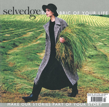 A woman in dark attire and a lengthy gray coat walks through a grassy field, holding a sizable bundle of green grass. A wide-brimmed black hat adorns her head. The text reads, "Selvedge - Issue 90: West by Selvedge: The Fabric of Your Life. Make Our Stories Part of Your Fashion Agenda.