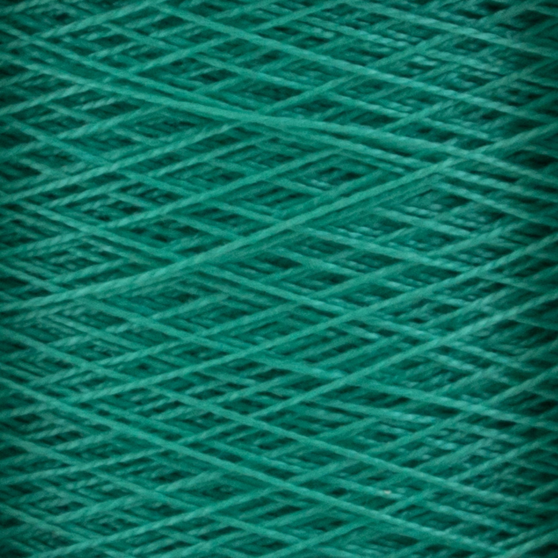 A close-up image of a spool of rich, vibrant green 10/2 Pearl Cotton Yarn from Supreme Corp. The threads are tightly wound around the mini-cone, creating a crisscross pattern. The texture is slightly glossy, giving it a subtle sheen. Renowned for its colorfastness, this versatile yarn ensures your projects remain stunning and enduring.