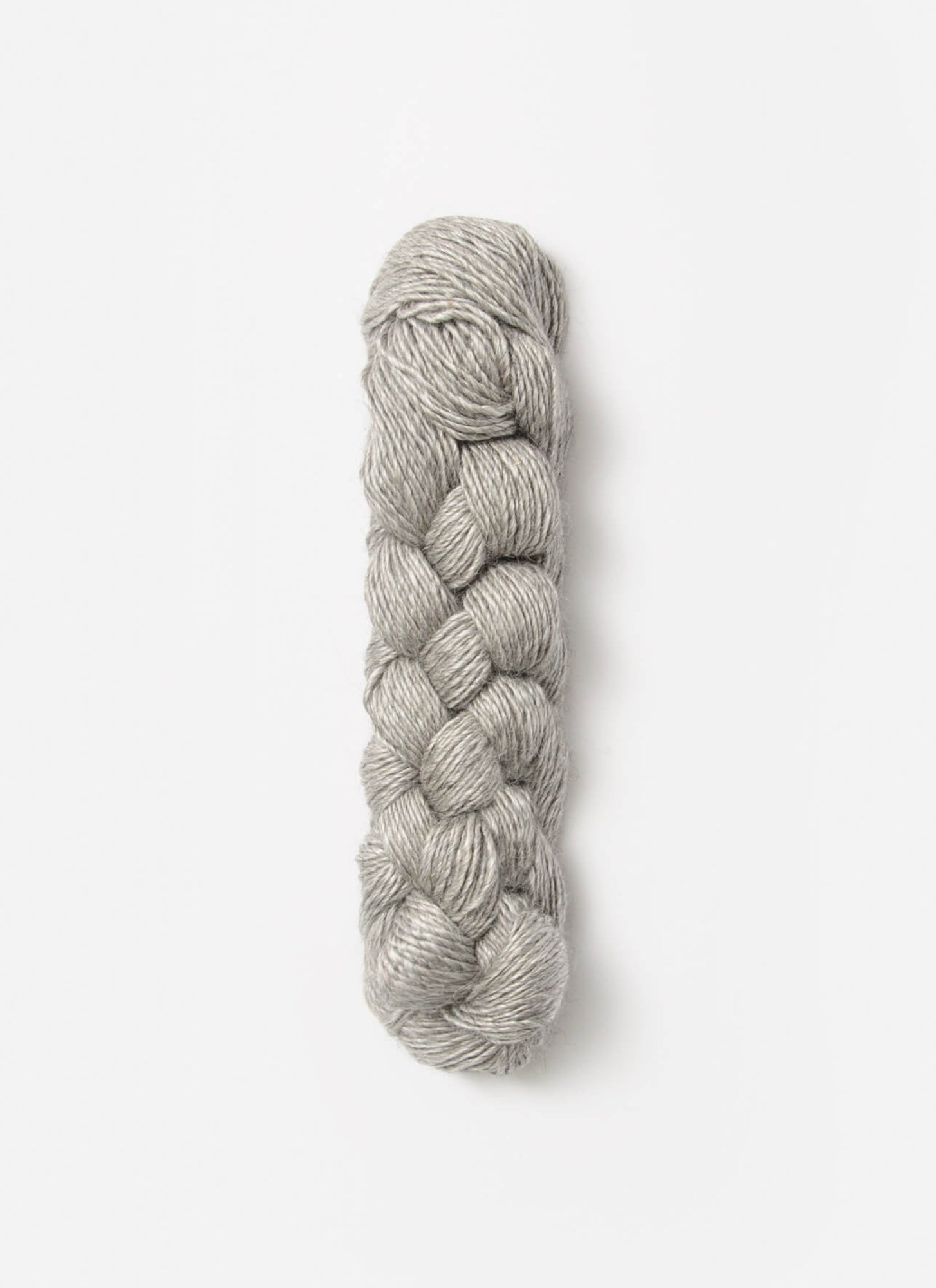 A neatly braided skein of light gray Blue Sky Fibers Metalico yarn against a plain white background. The silk blend yarn appears soft and has a subtle sheen, indicating a smooth texture. The braid is compact and evenly plaited, showcasing the quality and consistency of the sport weight fibers from Blue Sky Fibers.