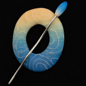 The Ring Ring Shawl Pin by Bonnie Bishoff Designs from Bonnie Bishoff is a lightweight, circular brooch featuring gradient colors that fade from peach at the top to blue at the bottom, adorned with white swirling patterns. Perfect for shawl closures, it includes a straight, slender metal pin with a matching gradient head running diagonally through the exquisite polymer clay design.