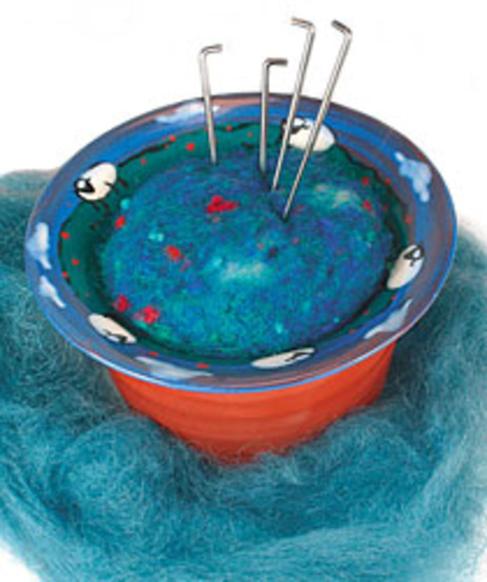 The Felting Needle Holder by Kick the Moon Farm is a colorful pin cushion shaped like a blue hill with red and green spots, nestled inside a decorated orange pot. This handmade needle holder features several metal hooks pinned into it, and is surrounded by a fluffy blue wool-like material that enhances its charm.