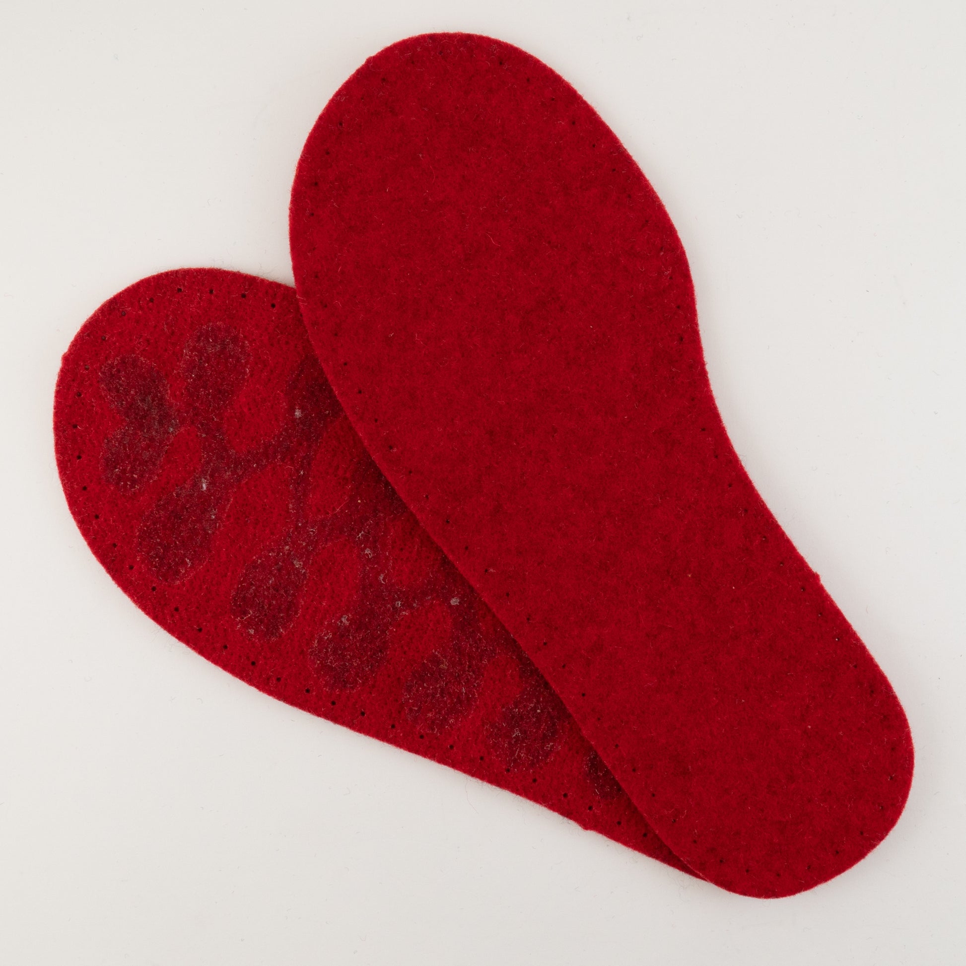 The Adult's Thick Felt Slipper Soles w/Latex Grip from Joe's Toes are featured against a white background. This pair of red felt-like wool insoles, resembling the sole of a felt slipper, are arranged with one insole overlapping the other. The back side reveals a non-slip surface with rubberized dots, making them ideal for any knit project or footwear.