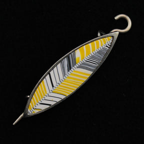 The Songbird Shawl Pin by Bonnie Bishoff Designs is an elongated oval brooch with a geometric design that boasts alternating black, white, and yellow stripes in a chevron-like pattern. This handmade accessory from Bonnie Bishoff features a metal border and a locking pin at one end for secure attachment, all set against a black background.