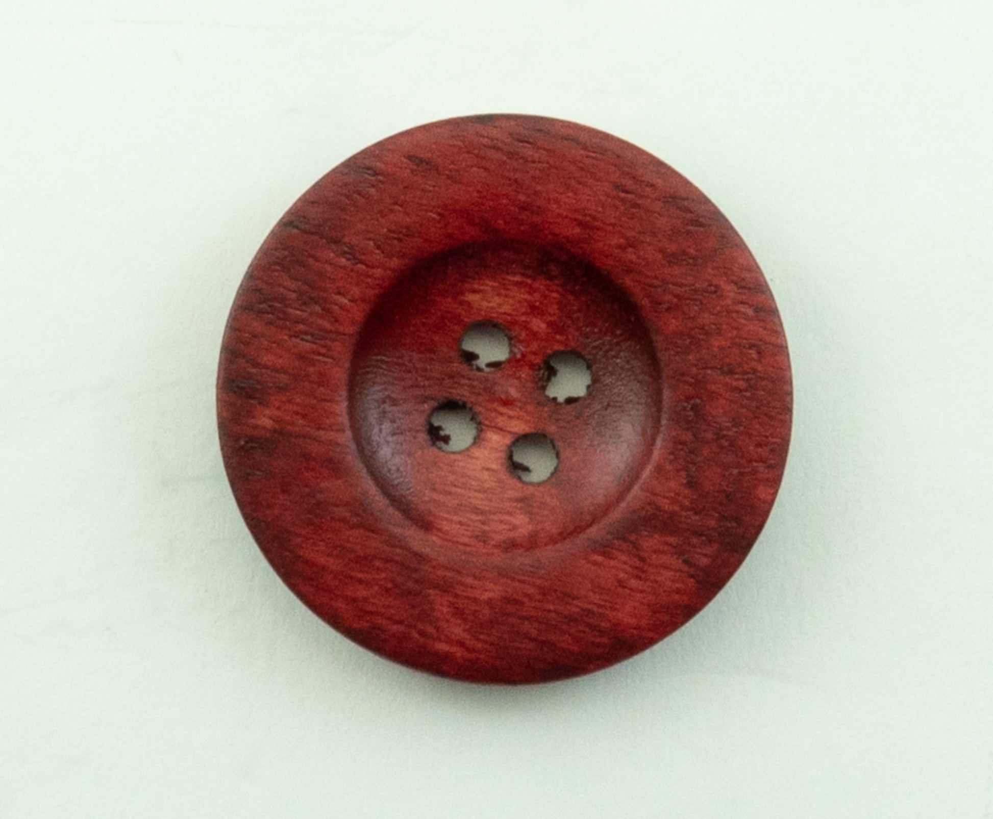 The Red Wood Saucer Button by Buttons Etc. is a round, wooden button with a deep reddish-brown finish. It features four holes in the center for sewing and has a diameter of 1 1/2". The visible wood grain adds texture to its polished surface, and it is placed on a white background.