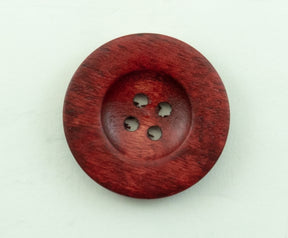 The Red Wood Saucer Button by Buttons Etc. is a round, wooden button with a deep reddish-brown finish. It features four holes in the center for sewing and has a diameter of 1 1/2". The visible wood grain adds texture to its polished surface, and it is placed on a white background.