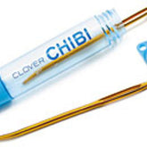 A blue plastic case with "Chibi Jumbo Darning/Tapestry Curved Needles" written on it, from Bryson Distributing, Inc., containing a gold-colored, easy-to-thread needle with a slight curve. The cap of the case is off, lying next to it. Another similar needle rests below the case. Perfect for sweater seaming.