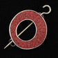 The Single Circle Shawl Pin by Bonnie Bishoff Designs expertly crafted by Bonnie Bishoff is a captivating brooch featuring a mesmerizing mosaic design in shades of red and black. Ideal for adorning your knit wearables, this circular accessory boasts an open center and is fastened with a silver pin that horizontally traverses the middle, complete with a hook on one end and a point on the other. The elegant design stands out against its black background.