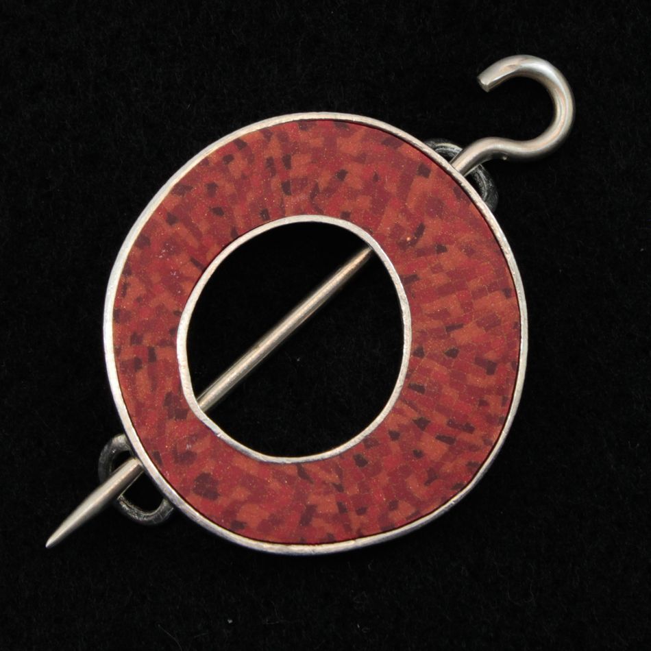 The Single Circle Shawl Pin by Bonnie Bishoff Designs expertly crafted by Bonnie Bishoff is a captivating brooch featuring a mesmerizing mosaic design in shades of red and black. Ideal for adorning your knit wearables, this circular accessory boasts an open center and is fastened with a silver pin that horizontally traverses the middle, complete with a hook on one end and a point on the other. The elegant design stands out against its black background.