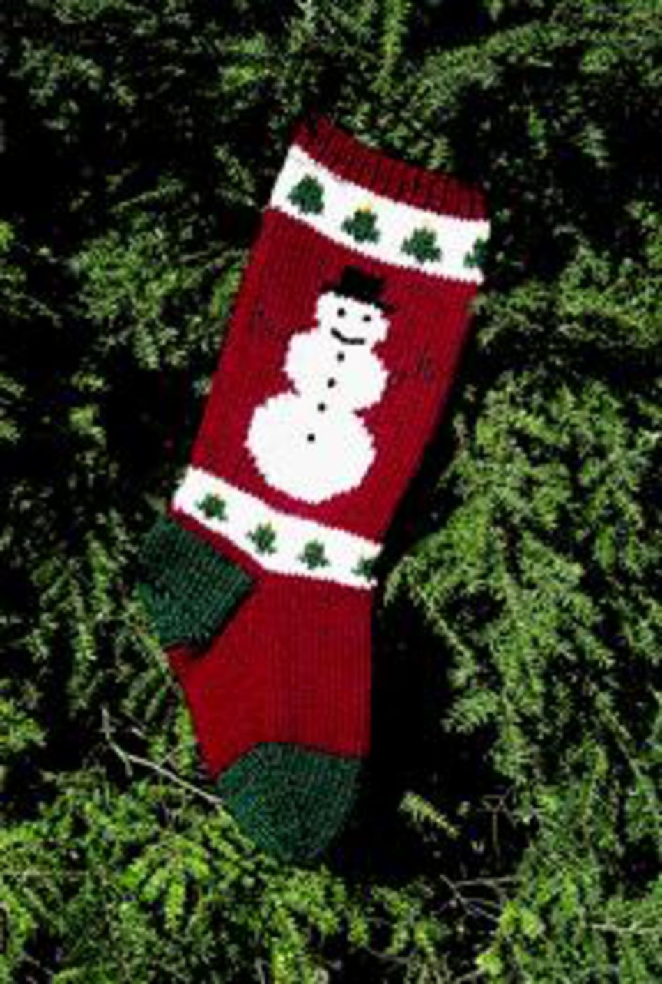 The Maine Christmas Stocking Kit from Bully Woolies is a festive stocking crafted from Maine spun wool. It features a charming white snowman design wearing a black top hat, set against a red background with green toe and heel sections. Additionally, the stocking is adorned with white bands showcasing green tree motifs, all beautifully placed on a backdrop of lush green foliage.