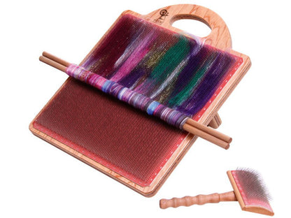 The Ashford Blending Board by Ashford Handicrafts Limited features a wooden design with a mix of colorful wool fibers partially rolled into rolags onto two wooden dowels. Beside the board is a small, wooden-handled brush used for blending the fibers. The board also includes an ergonomic handle at the top for easy use.