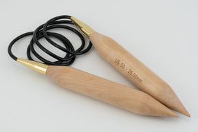 The Basix Jumbo Circular Knitting Needles by Accessories Unlimited are a pair of large circular wooden needles, ideal for jumbo yarns, connected by a flexible black cord. These needles feature smooth, tapered points and gold-colored metal connectors where they join the cord. One needle is engraved with "US 50 - 25.00 mm." The setup is displayed on a plain white background.