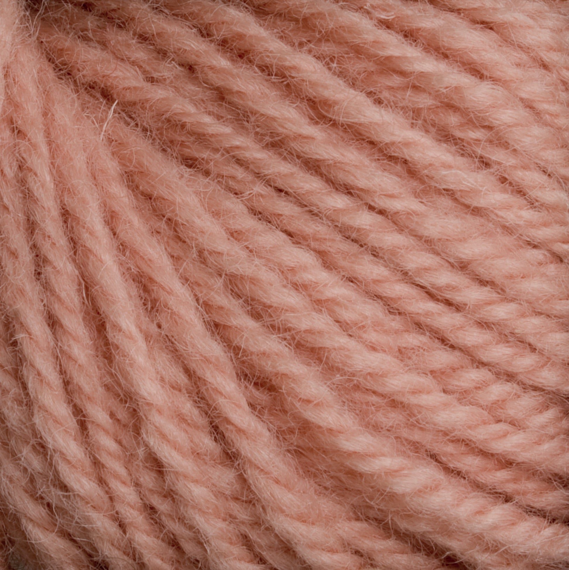 Close-up image of a skein of Halcyon Deco Rug Wool by Caledonian Dye Works. The soft, pinkish-peach yarn is tightly wound, showcasing its slightly fuzzy texture and twisted fibers. Made from 100% wool, the color is subtle and warm, making it ideal for cozy, handmade projects with versatile yarn options.