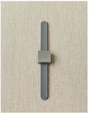 A minimalist grey wristwatch from Cocoknits, with a rectangular silver face and rubber strap, lies centered on a textured beige fabric background. The watch and strap have a simple, sleek design, evocative of the Maker's Keep Magnetic Bracelet aesthetic.