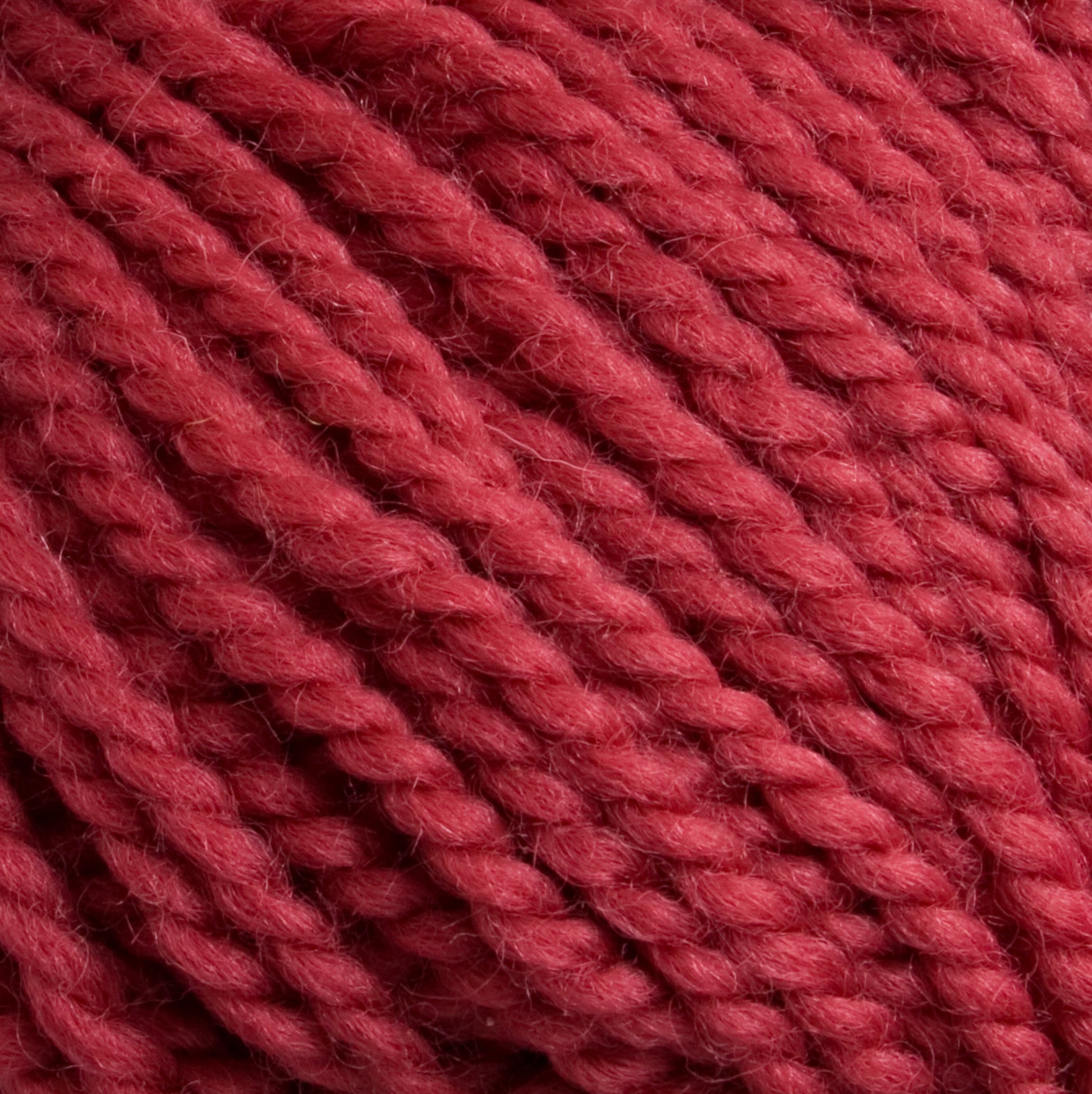 Close-up of a textured, thick red yarn. Norumbega by Caledonian Dye Works is an Aran weight yarn that is tightly twisted, creating a detailed, braided pattern. Made from 100% U.S. wool, the fibers appear soft and fluffy, highlighting the yarn’s cozy and warm characteristics suitable for knitting or crocheting.