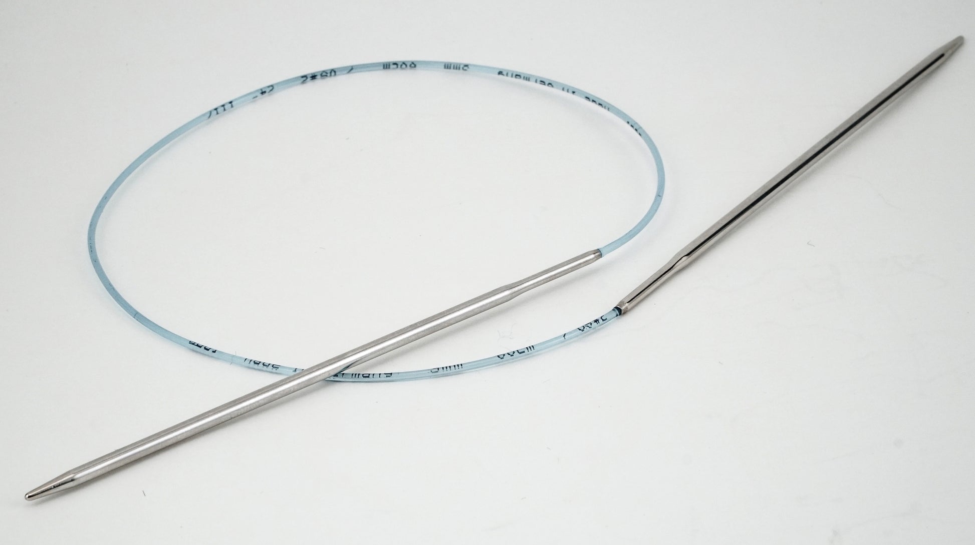 The image shows Addi Turbo Circular Knitting Needles from Skacel, featuring a flexible, thin wire coated in blue and coiled in a circle, resembling short circular needles. These knitting needles are connected to two rigid, straight metal ends and are used to guide yarn during knitting projects.