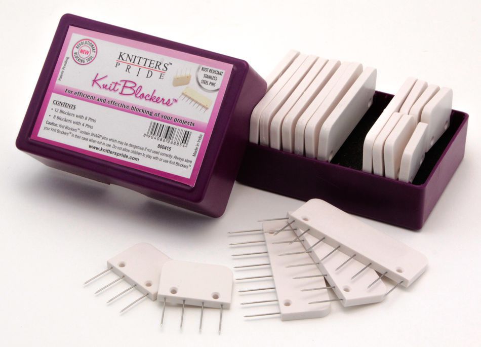 A purple box labeled "Knit Blockers by Knitter's Pride" from Accessories Unlimited sits open, revealing rows of white blockers with multiple pins. Additional knit blockers are arranged on blocking mats in front of the box, and the contents description on the label is partially visible.