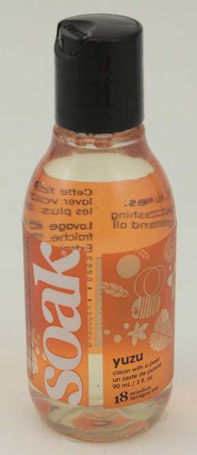 A clear plastic bottle with a black flip-top cap contains Soak Wash brand liquid detergent in the Yuzu scent. The biodegradable label is orange with white text, featuring patterns of leaves and flowers. Suitable for delicate wash items, this no-rinse soap from Soak Wash Inc. comes in a 90 mL size, sufficient for 18 washes.