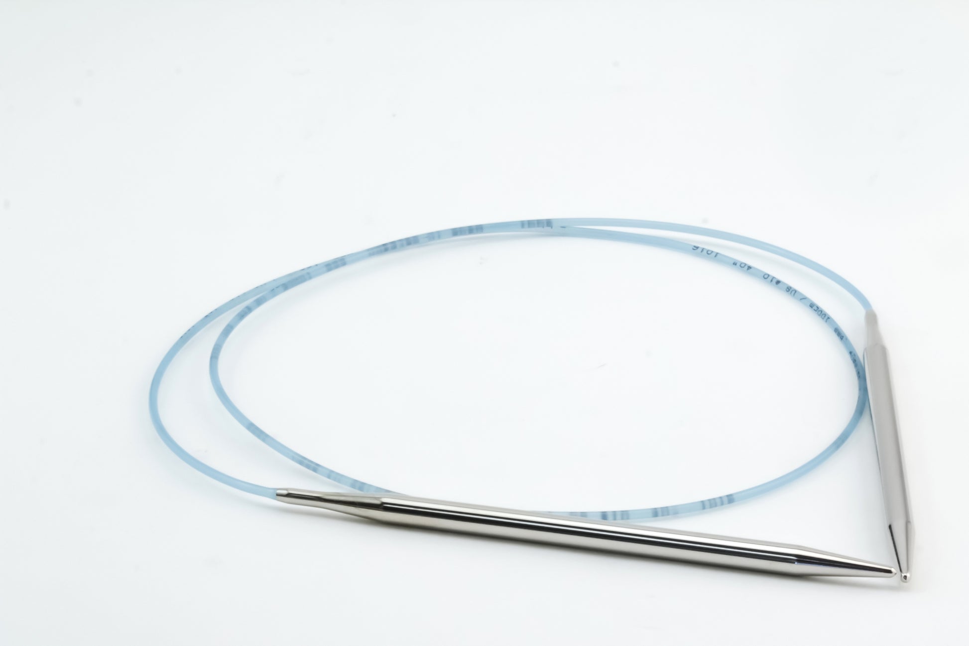 A pair of Addi Turbo Circular Knitting Needles from Skacel lies on a white surface, connected by a flexible blue cable. The short circular needles have pointed tips on both ends, and the cable forms a loop. Marked with measurements, this setup is perfect for crafting cuffs, collars, or doll clothes.