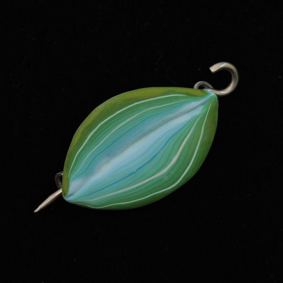 The Hosta Shawl Pin by Bonnie Bishoff Designs is a creation by Bonnie Bishoff that highlights the intricate beauty of polymer clay art. Shaped like a green leaf, this pin features a metallic hook on one end and a loop on the other. White and light blue lines run through the center, forming a striped pattern against a black background.