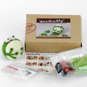 Photo of a Woolbuddy Needle Felting Kit. The kit features a felted frog with big eyes, a branded Woolbuddy cardboard box, detailed instructions, and bags containing colored wool roving and felting needles.