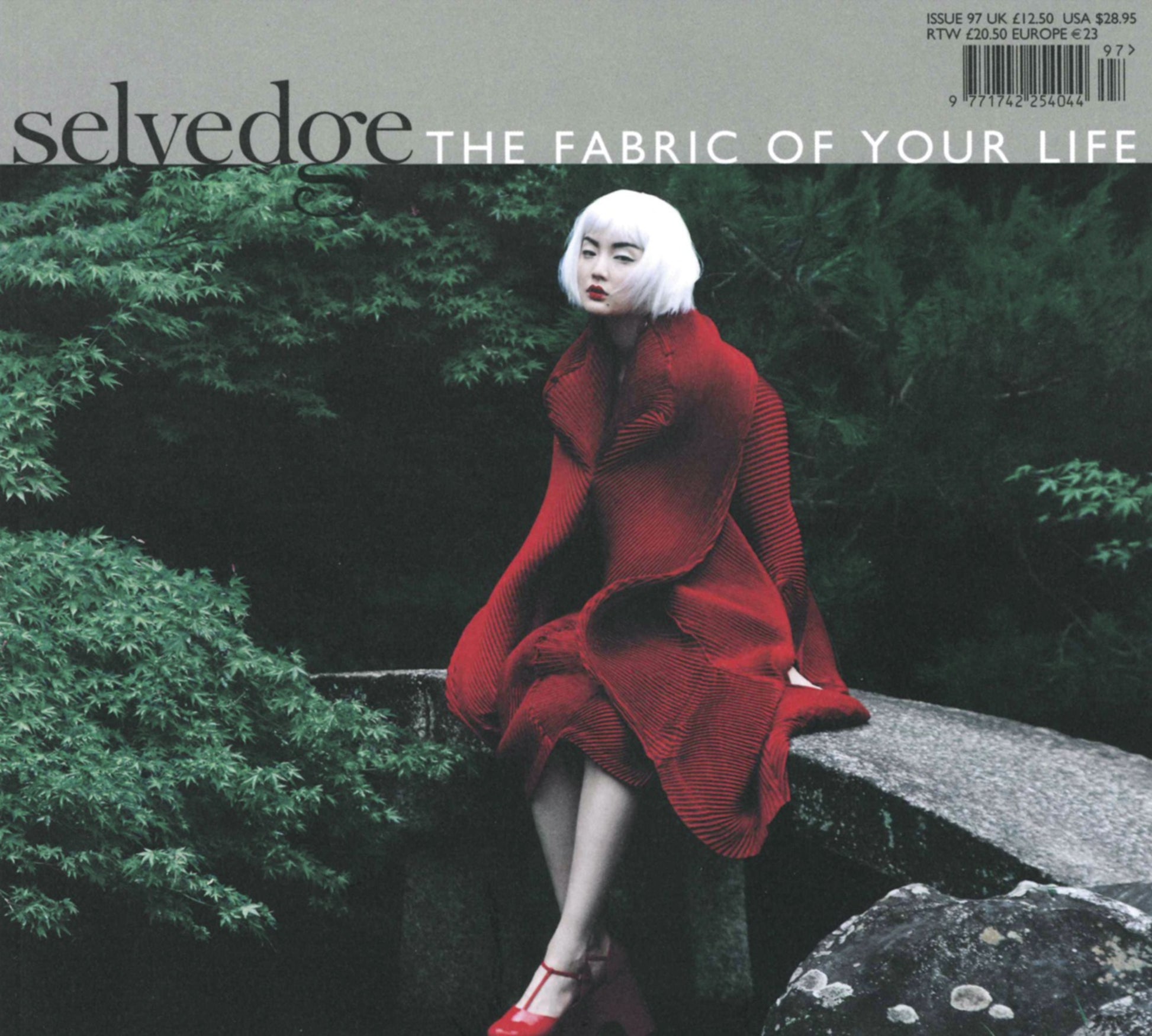 A woman with short white hair, dressed in a vibrant red outfit and matching red shoes, sits on a stone ledge outdoors surrounded by lush green foliage. The cover text of Selvedge - Issue 97: Red reads "Selvedge: The Fabric of Your Life." At the top right, the price and issue details celebrate textiles in fashion.