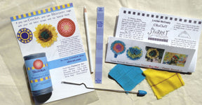 Laid out on a light surface is the Shirret Everything Kit from Shirret. Items include crochet yarn, a special hook, fabric strips, crochet directions, and a pencil. The kit features colorful patterns and designs for creating a crocheted flower in vibrant blue and yellow tones.